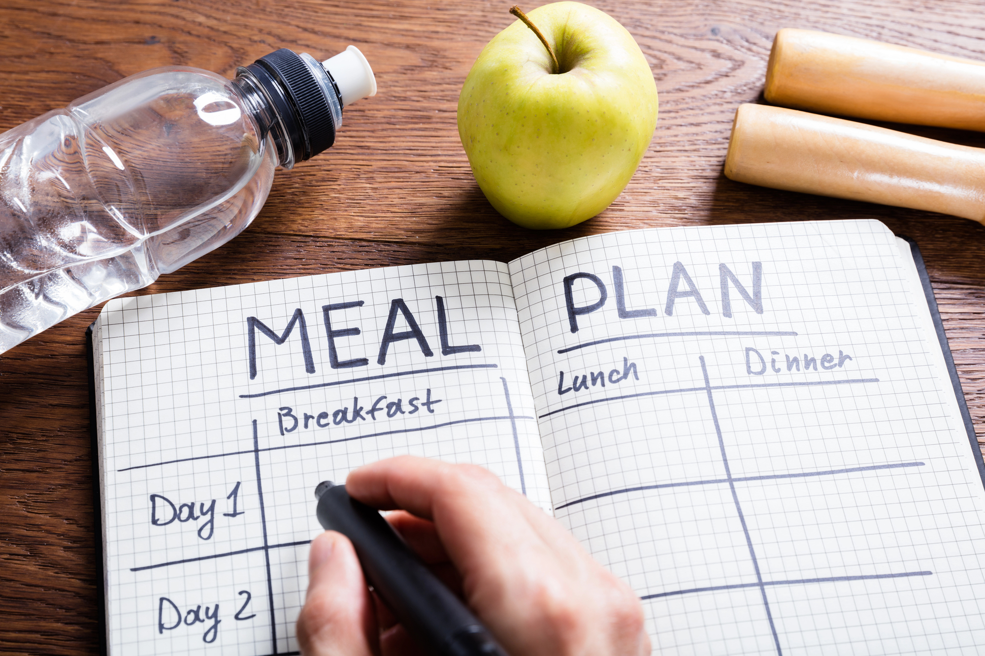 A hand writes in a notebook labeled "Meal Plan" with sections for breakfast, lunch, and dinner. Next to the notebook are a filled water bottle, a green apple, and two beige dumbbells on a wooden surface.