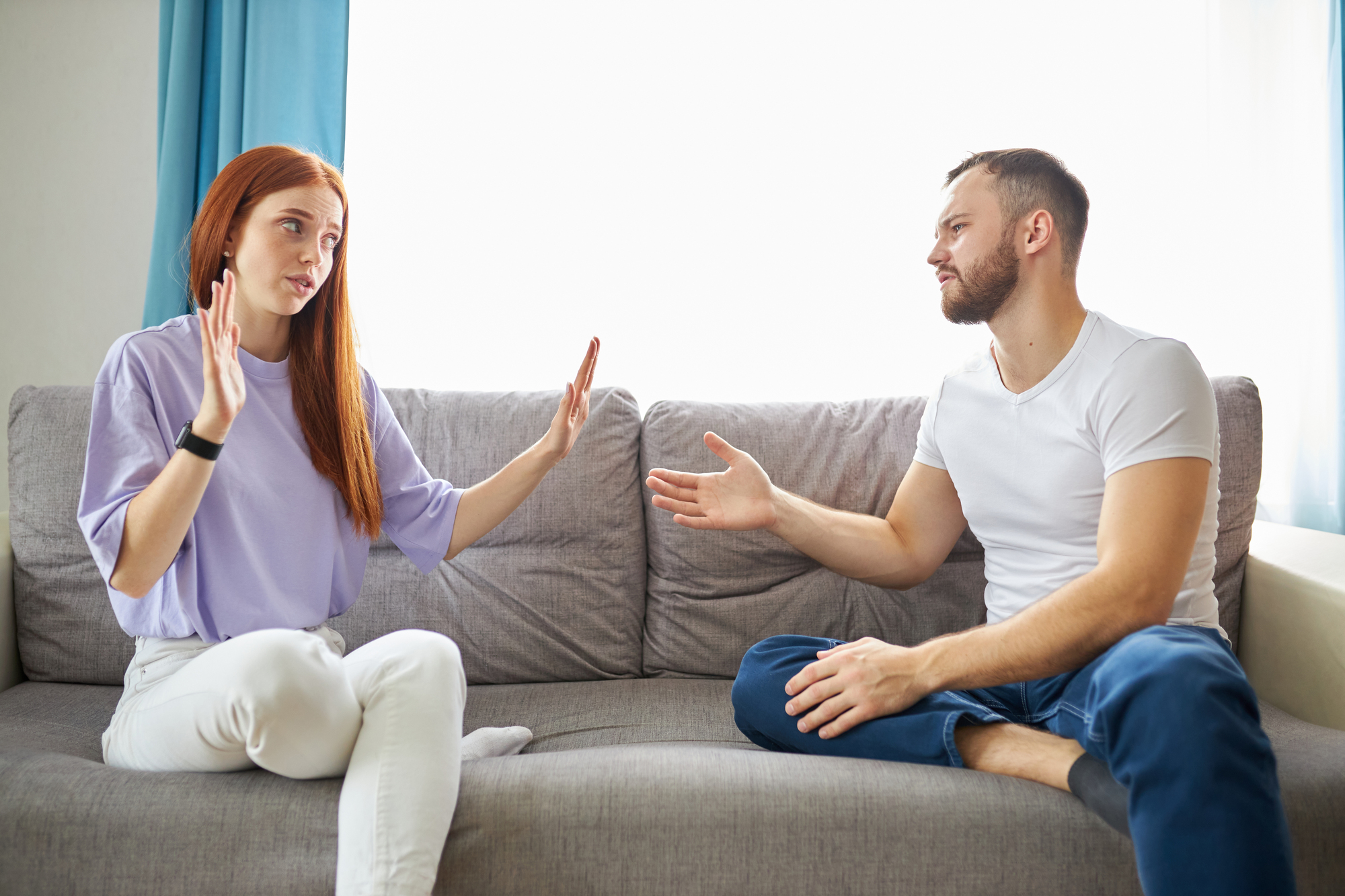 A woman with long red hair, wearing a light purple top and white pants, sits on a gray couch raising her hands defensively while talking to a man with a beard, wearing a white t-shirt and jeans, who is extending his hand towards her. They appear to be in a heated discussion.