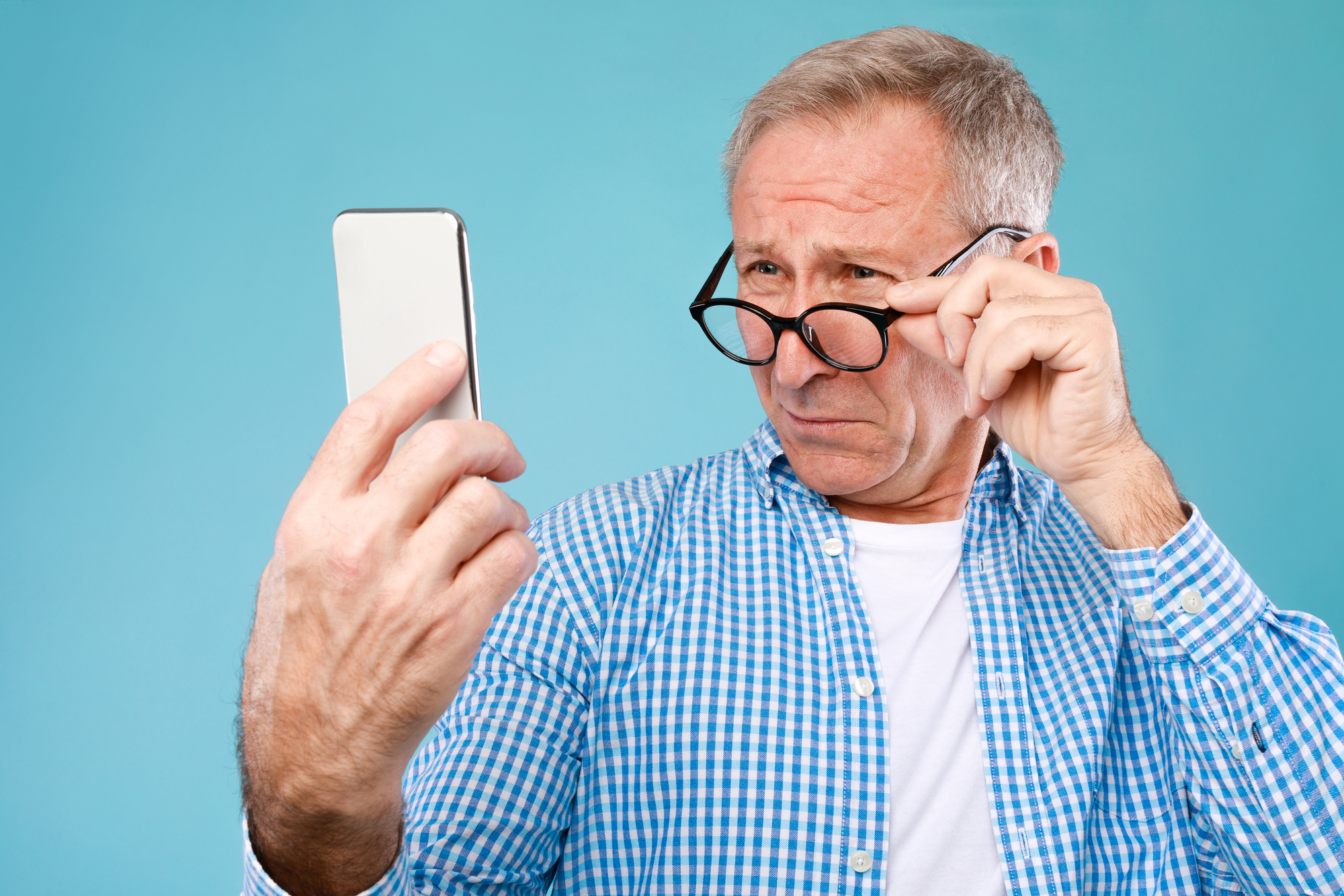 An older man in a blue and white checkered shirt holds a smartphone at arm's length while adjusting his glasses with a puzzled expression. He appears to be trying to read the screen. The background is a solid light blue.