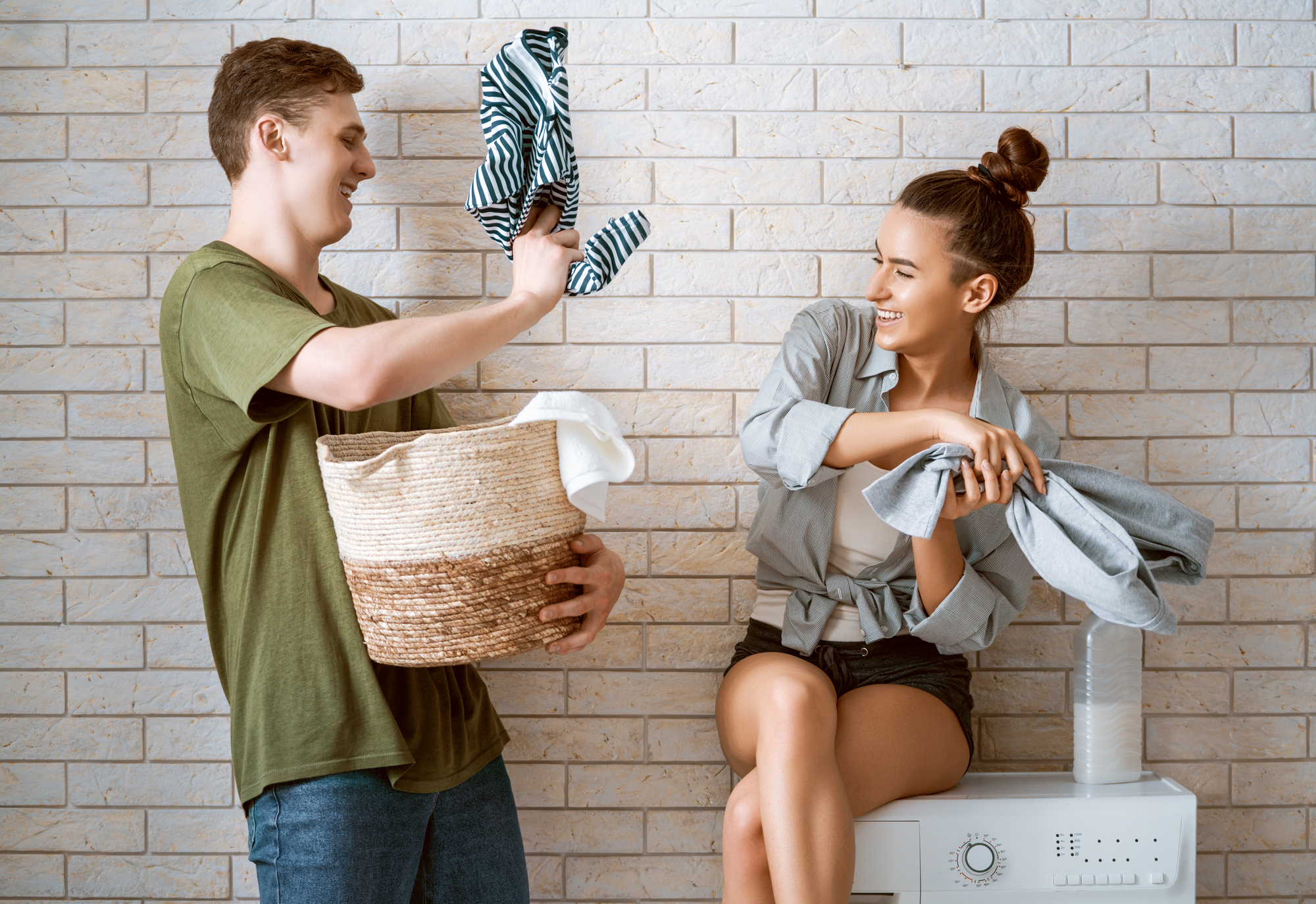 A young man and woman are doing laundry together. The man is jokingly holding a shirt towards the woman, while she sits on a washing machine holding clothes. They both laugh and smile in a light, playful moment against a tiled wall background.