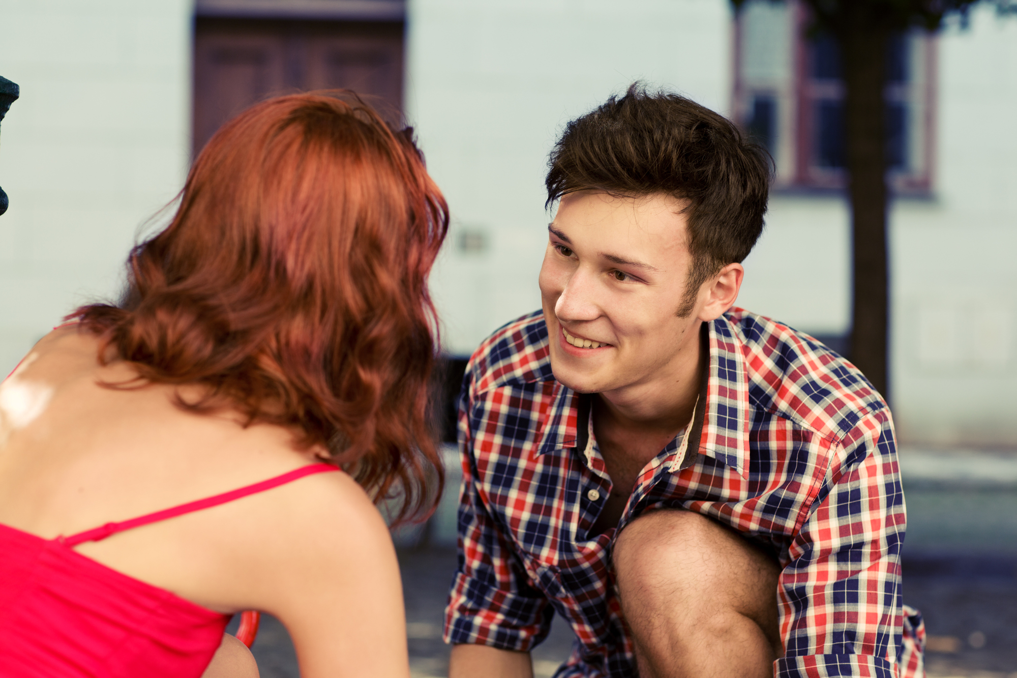 A young man with short dark hair, wearing a plaid shirt, smiles at a woman with red hair in a red dress. They are outdoors, with a blurred building and trees in the background.