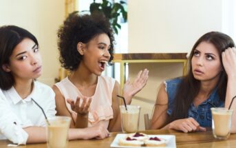 Three women sitting at a table with drinks and desserts. The woman on the left appears indifferent, the woman in the middle is animatedly talking, and the woman on the right looks annoyed, resting her head on her hand.