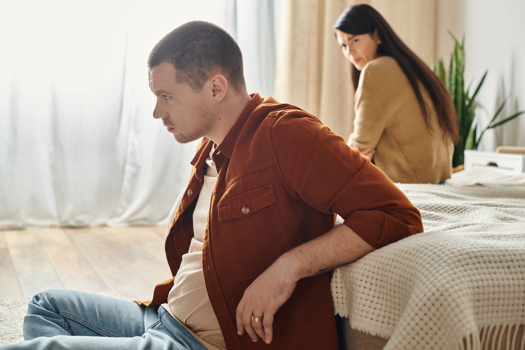 A man in a rust-colored jacket sits on the floor with a solemn expression, leaning against a bed. In the background, a woman in a tan sweater sits on the bed, looking at him with concern. The room has wooden floors, light curtains, and a plant in the corner.