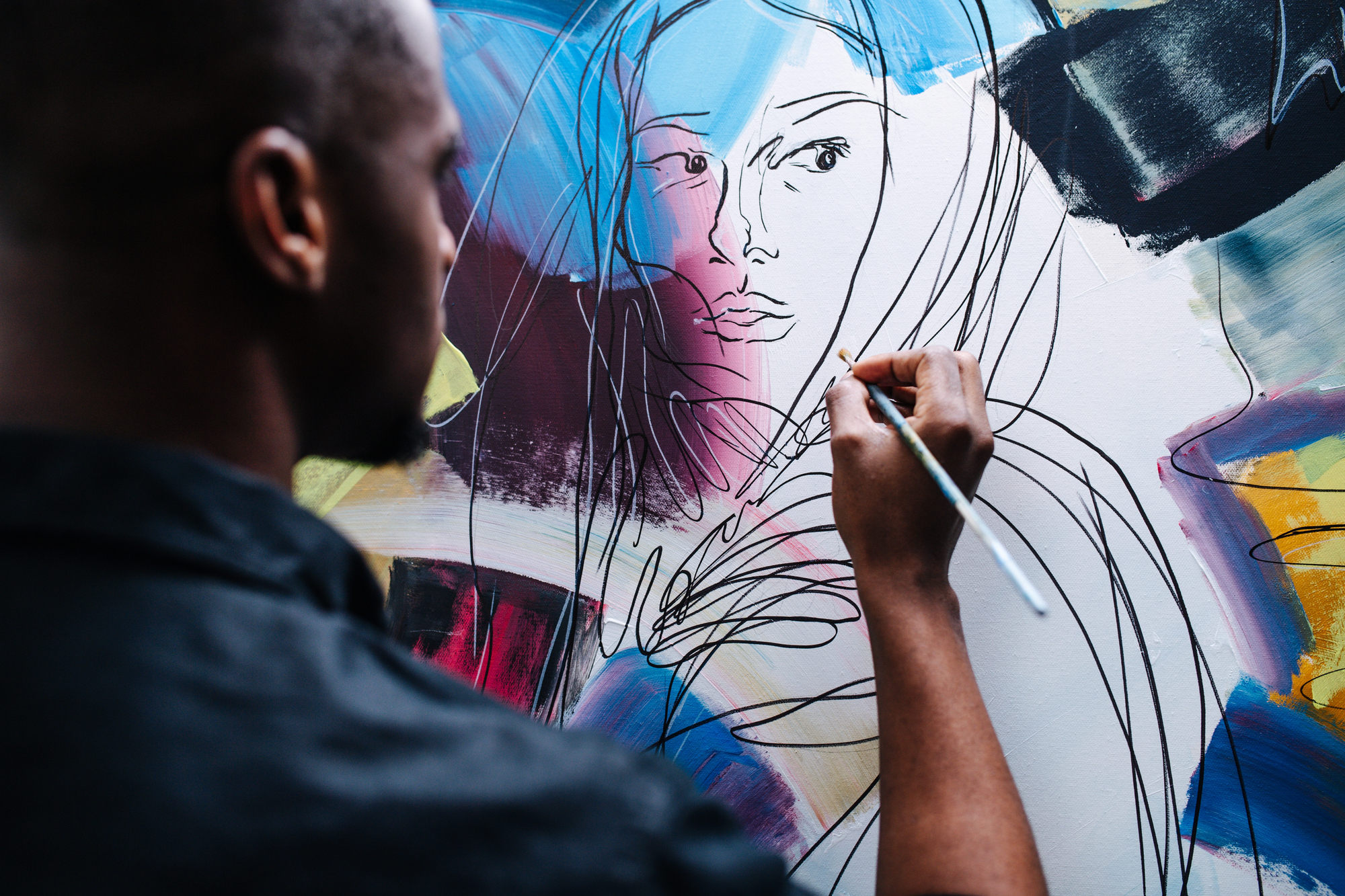 A person with short hair is painting on a canvas. The artwork appears to be an abstract depiction of a woman, featuring bold lines and vibrant colors. The artist is using a small brush and focusing on the details of the face.