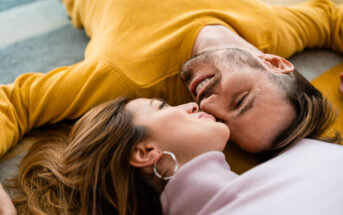 A couple lies on the floor, their heads close together. The man, wearing a yellow sweater, looks lovingly at the woman, who wears a lavender top. Both appear relaxed and happy, sharing an intimate moment.