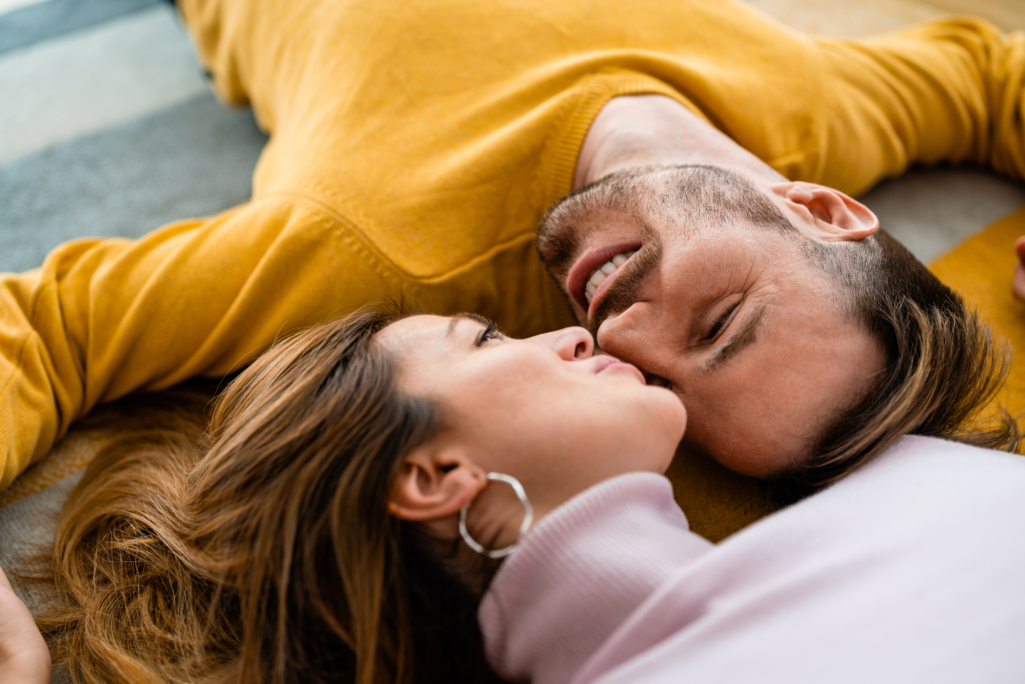 A couple lies on the floor, their heads close together. The man, wearing a yellow sweater, looks lovingly at the woman, who wears a lavender top. Both appear relaxed and happy, sharing an intimate moment.