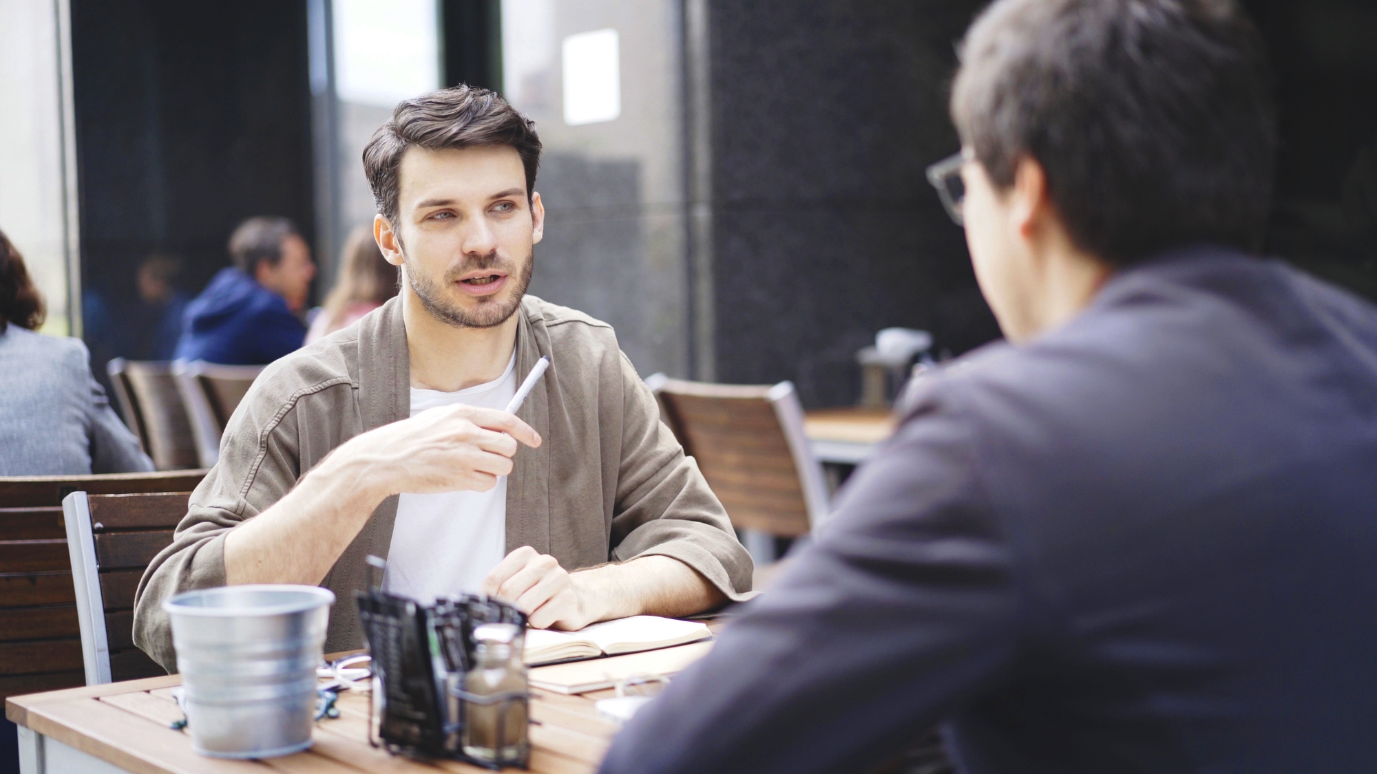 Two men are sitting at a wooden table in an outdoor café, engaged in a conversation. One man, holding a pen, is speaking, while the other listens attentively with his back to the camera. The table holds a metal bucket, condiments, and utensils. Other patrons are blurred in the background.