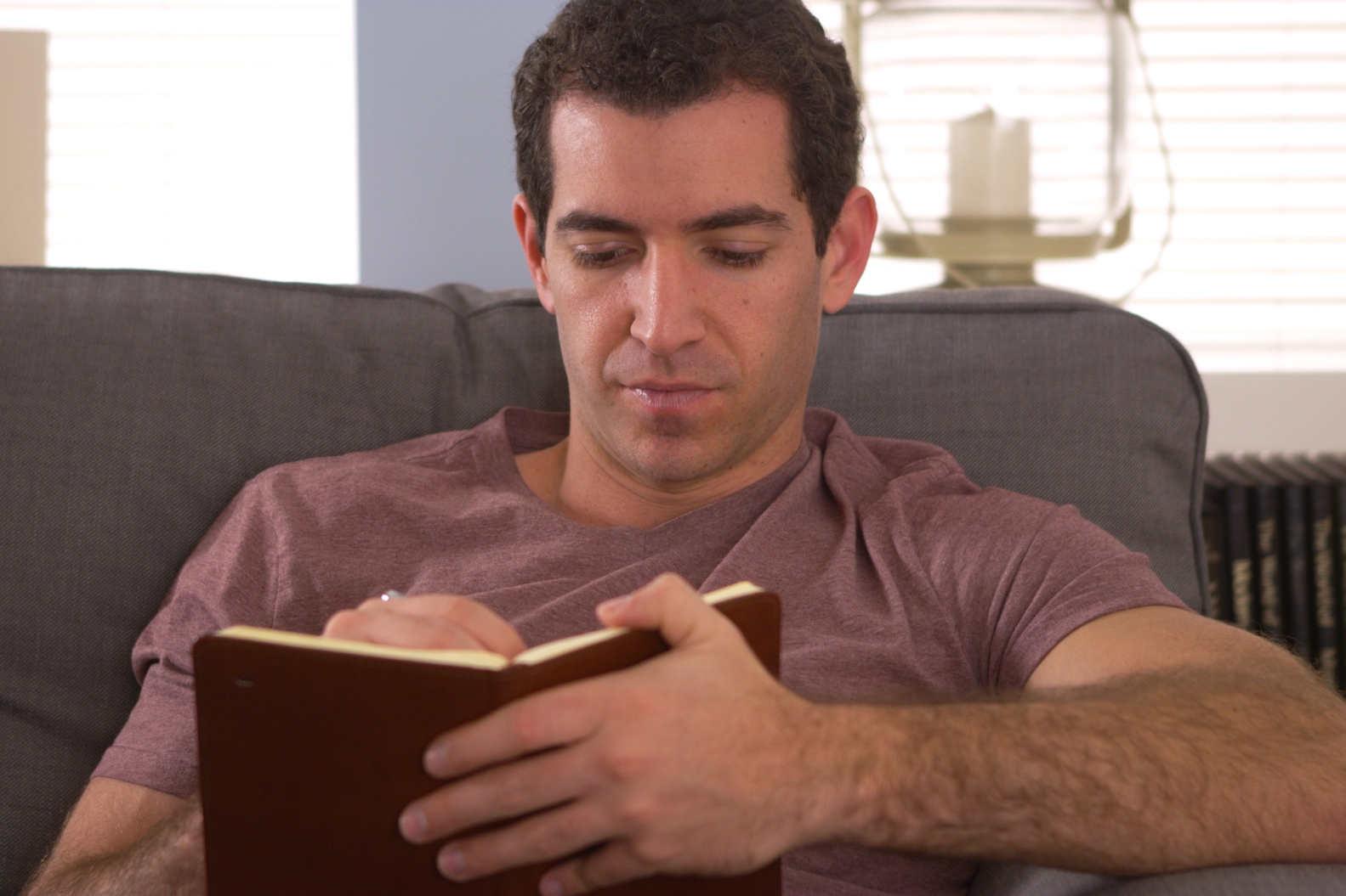 A person with short, curly hair and a maroon shirt is sitting on a couch, writing in a brown notebook. There is a bookshelf with black and yellow books and a lantern behind them. The scene appears calm and focused.