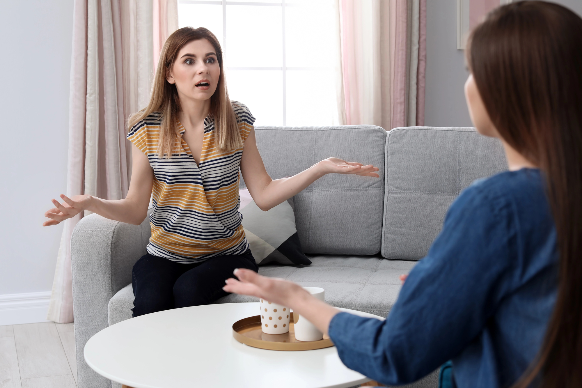 Two women sit on a light gray couch in a living room, engaged in a heated conversation. The woman on the left wears a striped shirt and has her hands raised, expressing frustration. The woman on the right, with long hair, faces her and also gestures while speaking.