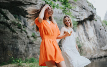 Two women are dancing joyfully outdoors near a rocky terrain. One is wearing a bright orange dress, while the other is dressed in white. They appear to be laughing and twirling, with their hair and dresses flowing in motion under the natural light.