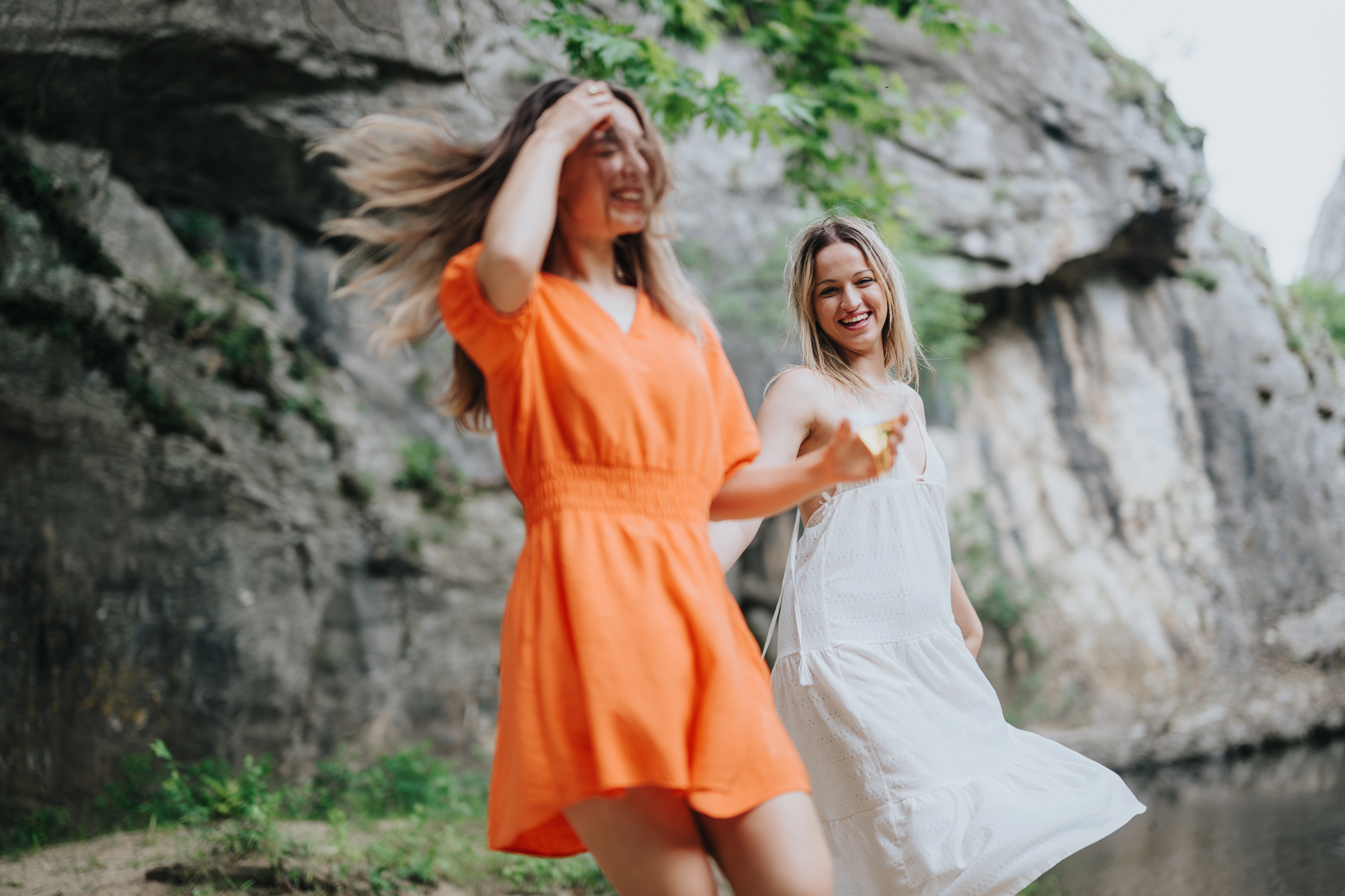 Two women are dancing joyfully outdoors near a rocky terrain. One is wearing a bright orange dress, while the other is dressed in white. They appear to be laughing and twirling, with their hair and dresses flowing in motion under the natural light.