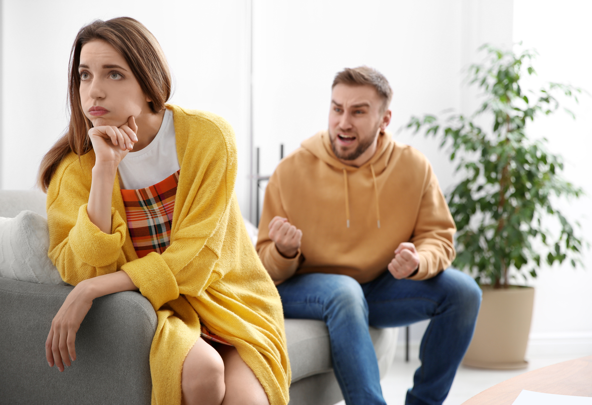 A woman in a yellow cardigan sits on a couch, looking away with a hand on her chin, appearing frustrated. A man in a brown hoodie sits next to her, gesturing with clenched fists and an angry expression, amidst a dispute in a bright room with plants.