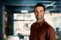 A man wearing a brown shirt smiles confidently in an office environment. The space is modern with large windows and a conference table in the background.