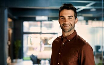 A man wearing a brown shirt smiles confidently in an office environment. The space is modern with large windows and a conference table in the background.