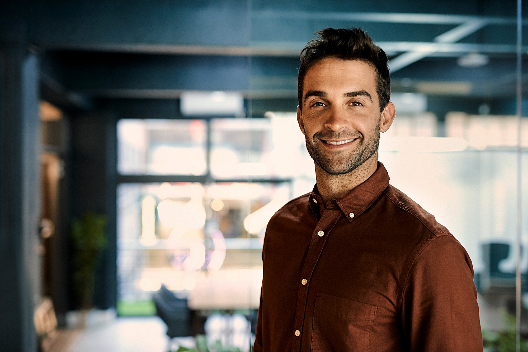 A man wearing a brown shirt smiles confidently in an office environment. The space is modern with large windows and a conference table in the background.