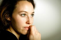 A woman with light eyes and freckles gazes thoughtfully to the right. Her hand is near her mouth, and she is wearing a black top. The background is a soft beige, creating a calm atmosphere.