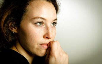 A woman with light eyes and freckles gazes thoughtfully to the right. Her hand is near her mouth, and she is wearing a black top. The background is a soft beige, creating a calm atmosphere.