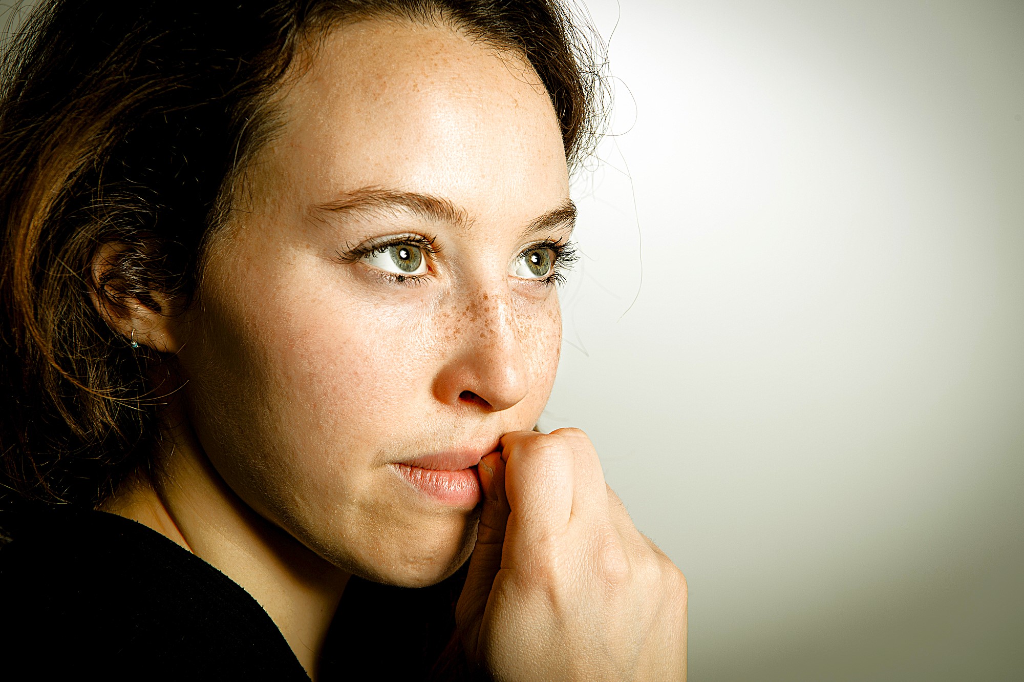 A woman with light eyes and freckles gazes thoughtfully to the right. Her hand is near her mouth, and she is wearing a black top. The background is a soft beige, creating a calm atmosphere.