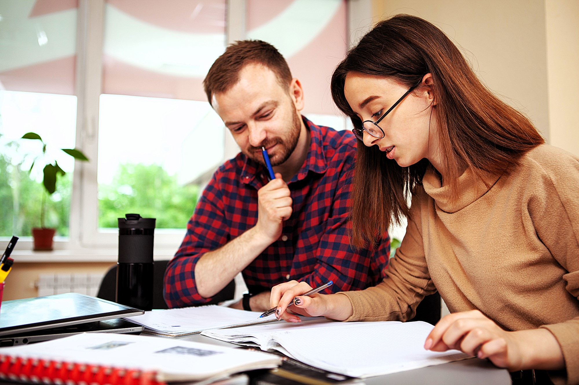 A man and a woman are sitting at a desk, focused on writing and reviewing documents. The woman is wearing glasses, and both have pens in hand. A laptop and notebooks are on the desk, with a plant visible in the background by a window.