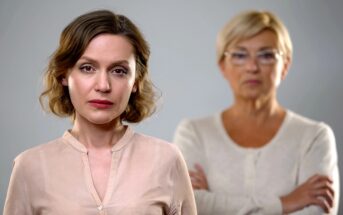 A woman with short brown hair and a neutral expression stands in the foreground. In the blurred background, an older woman with short blonde hair and glasses has her arms crossed. Both wear light-colored tops against a gray background.