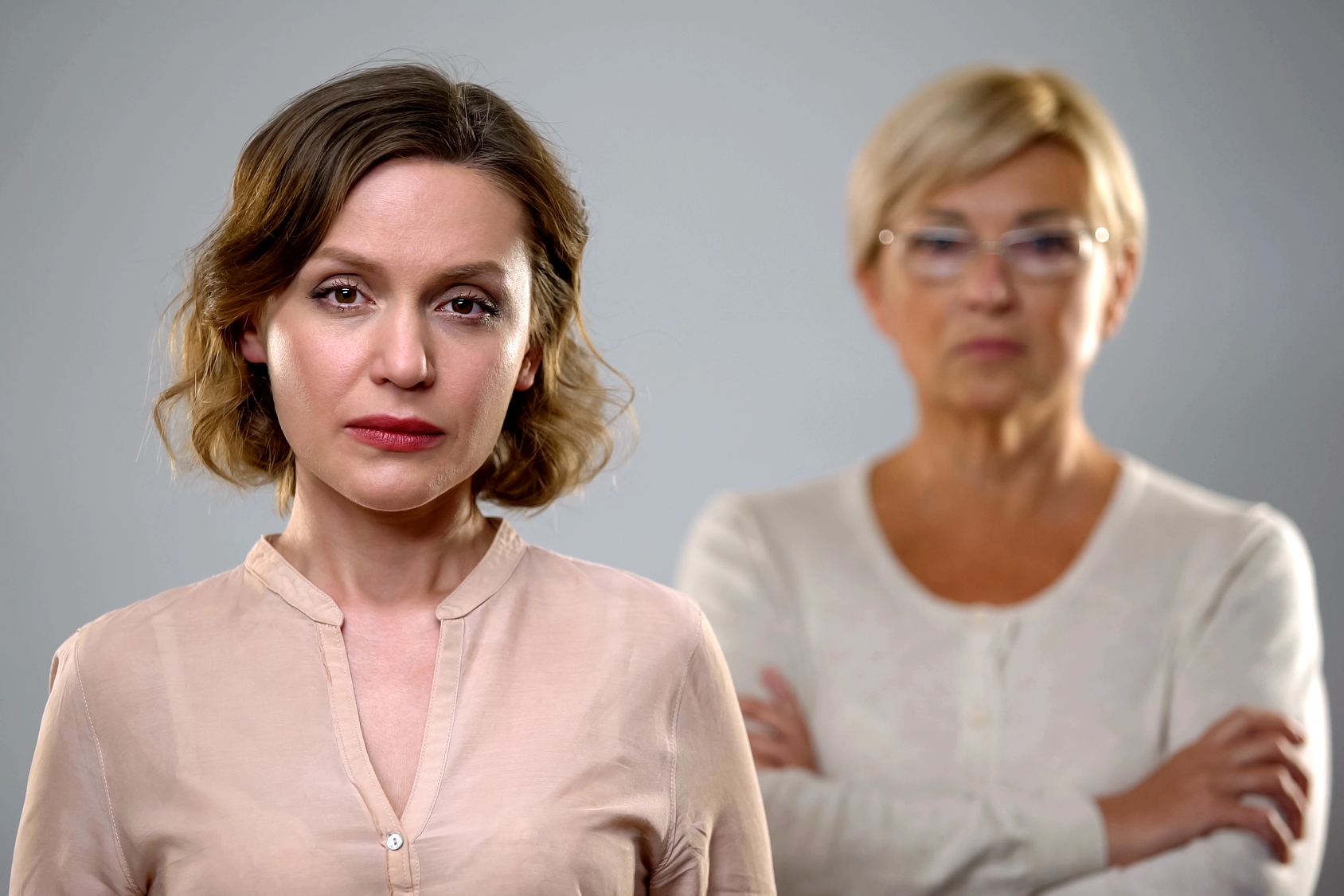 A woman with short brown hair and a neutral expression stands in the foreground. In the blurred background, an older woman with short blonde hair and glasses has her arms crossed. Both wear light-colored tops against a gray background.