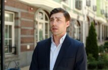 A man in a dark suit stands outdoors in front of a building with arched windows and a garden. He has short brown hair and a white shirt, and he appears to be talking or making a statement.