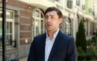 A man in a dark suit stands outdoors in front of a building with arched windows and a garden. He has short brown hair and a white shirt, and he appears to be talking or making a statement.