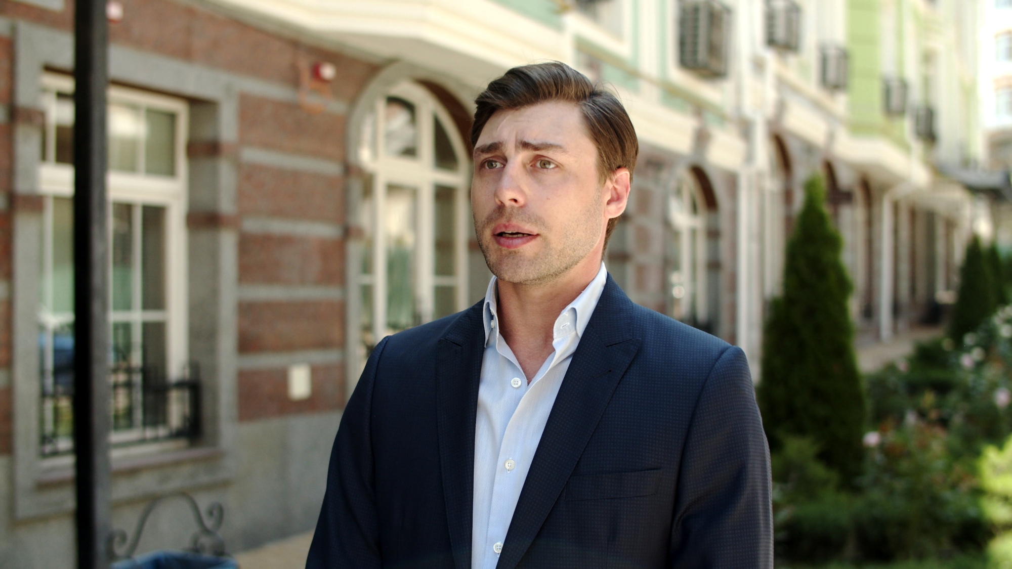 A man in a dark suit stands outdoors in front of a building with arched windows and a garden. He has short brown hair and a white shirt, and he appears to be talking or making a statement.