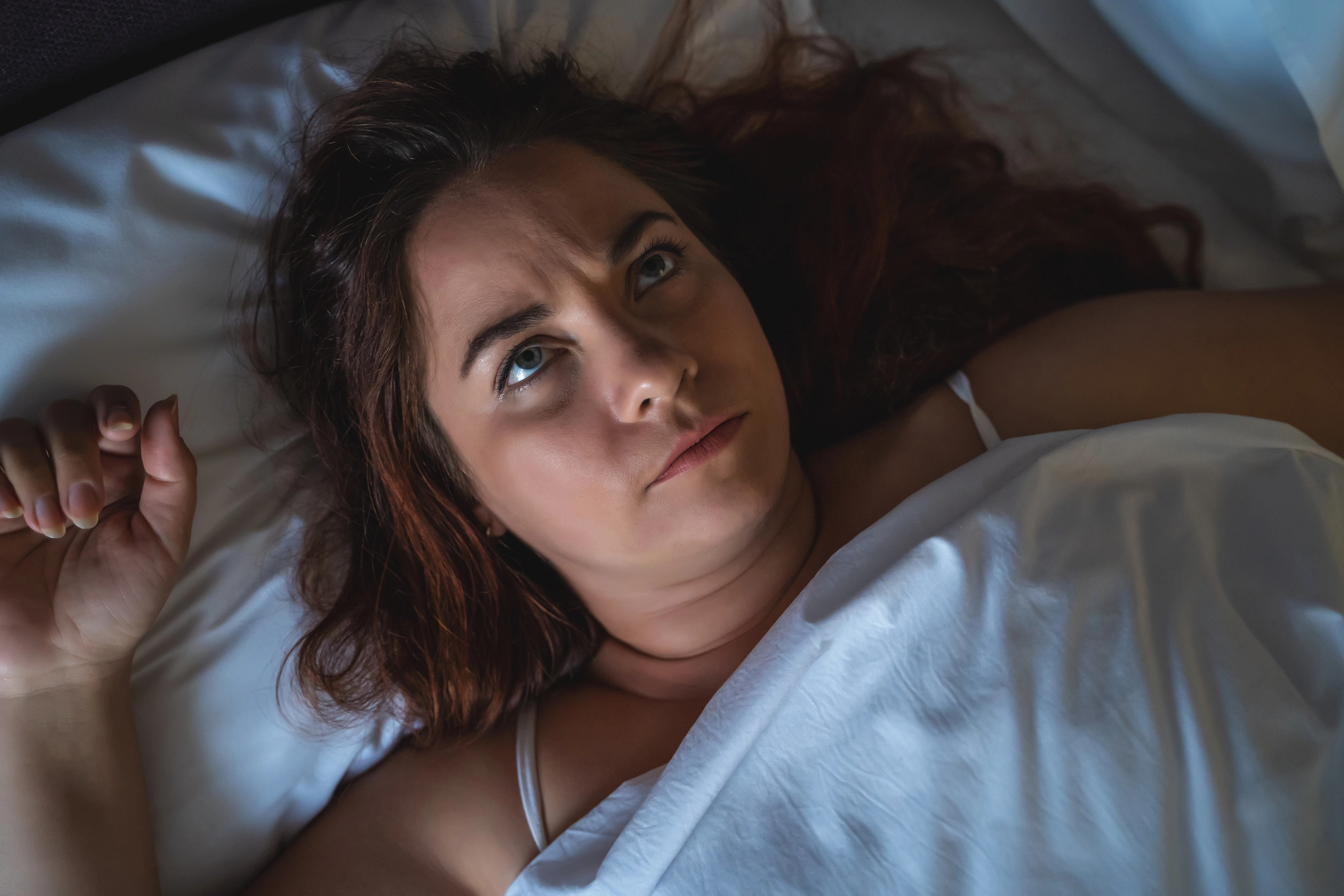 A woman with long dark hair is lying in bed, partially covered by a white sheet. She has a concerned or thoughtful expression on her face, looking up towards the ceiling. Her hand rests on the pillow next to her head.