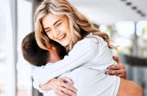 A woman with long blonde hair is joyfully hugging a man indoors. She is smiling broadly, and they both appear happy. The room is well-lit with natural light coming through large windows in the background.