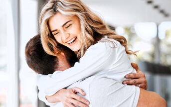 A woman with long blonde hair is joyfully hugging a man indoors. She is smiling broadly, and they both appear happy. The room is well-lit with natural light coming through large windows in the background.