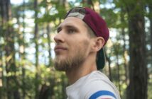 A man with a beard and a maroon baseball cap worn backwards gazes off into the distance amid a forest setting. He wears a white shirt with blue, red, and white stripes on the sleeve. The background features trees with green leaves.