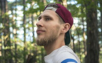 A man with a beard and a maroon baseball cap worn backwards gazes off into the distance amid a forest setting. He wears a white shirt with blue, red, and white stripes on the sleeve. The background features trees with green leaves.