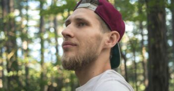 A man with a beard and a maroon baseball cap worn backwards gazes off into the distance amid a forest setting. He wears a white shirt with blue, red, and white stripes on the sleeve. The background features trees with green leaves.