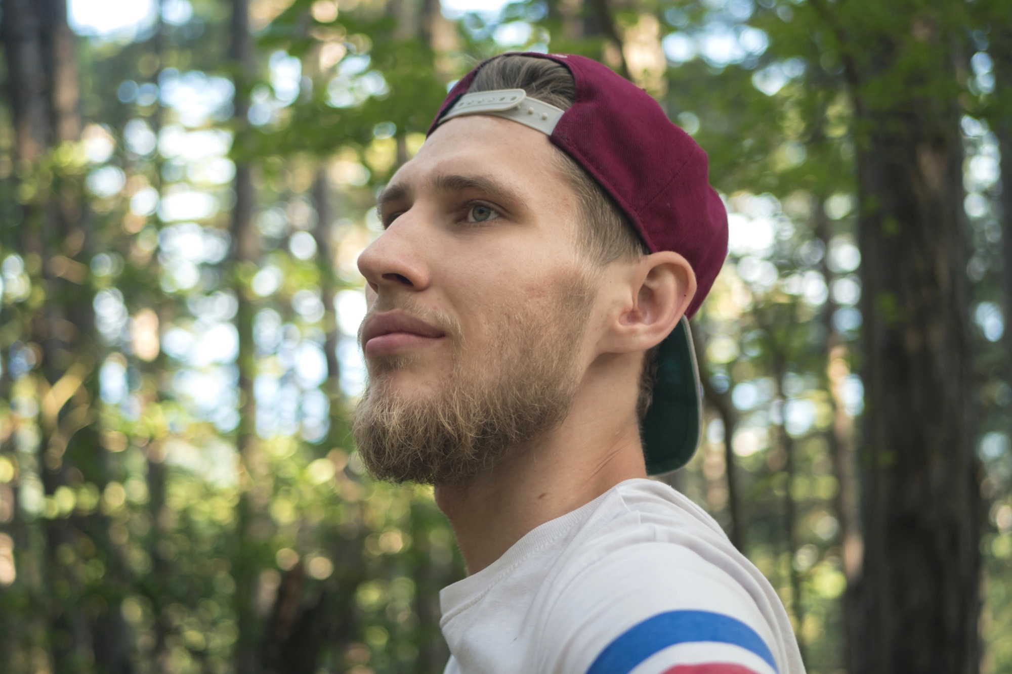 A man with a beard and a maroon baseball cap worn backwards gazes off into the distance amid a forest setting. He wears a white shirt with blue, red, and white stripes on the sleeve. The background features trees with green leaves.