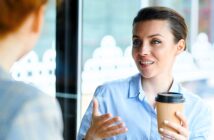 A woman in a blue shirt holds a coffee cup while engaging in conversation with someone off-camera. She appears animated and is using hand gestures. The background shows blurred windows with decorative patterns.