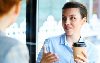 A woman in a blue shirt holds a coffee cup while engaging in conversation with someone off-camera. She appears animated and is using hand gestures. The background shows blurred windows with decorative patterns.