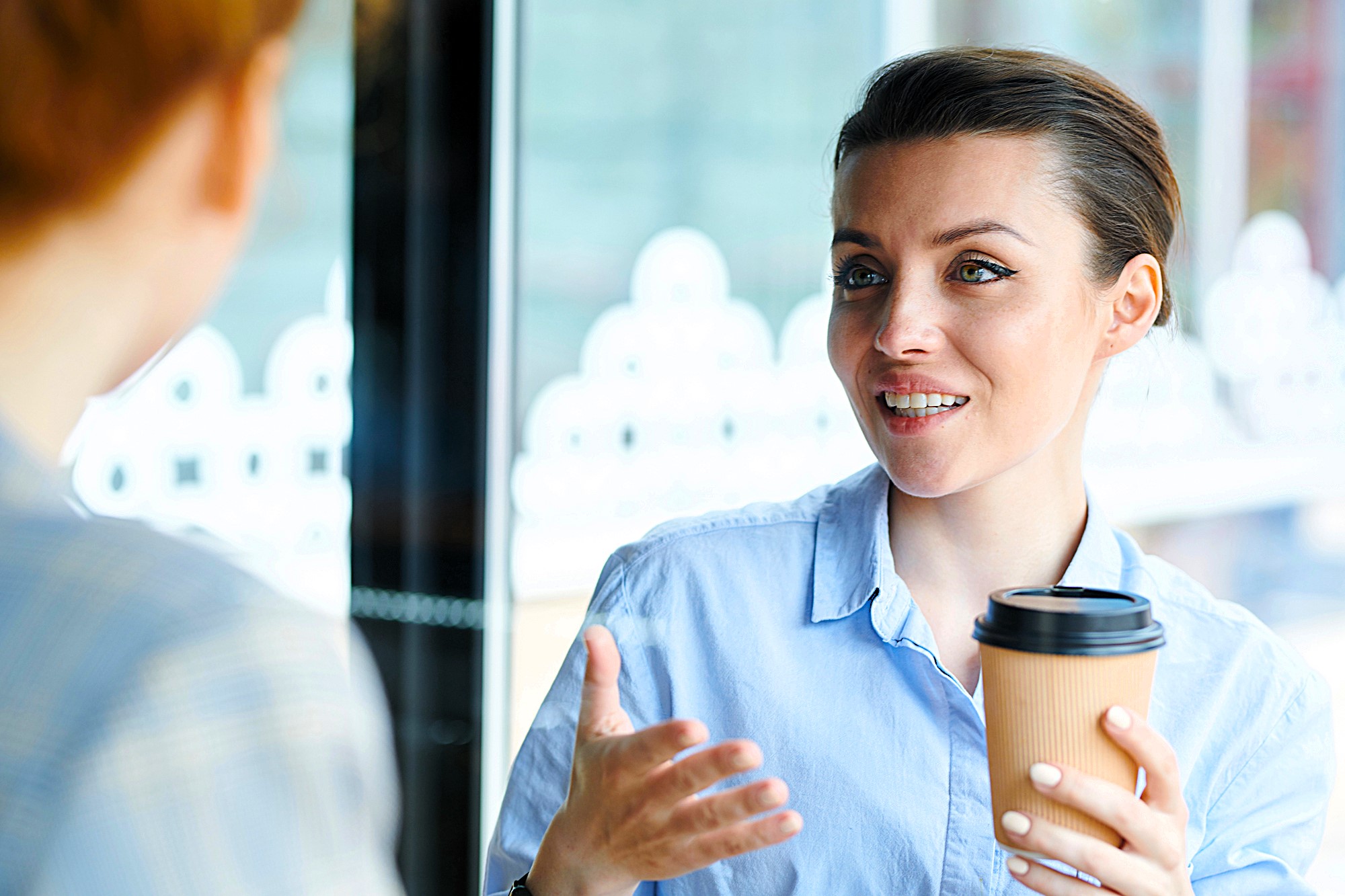 A woman in a blue shirt holds a coffee cup while engaging in conversation with someone off-camera. She appears animated and is using hand gestures. The background shows blurred windows with decorative patterns.