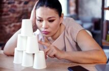 A woman rests her head on one hand while intently stacking paper cups into a pyramid on a desk. She appears focused, with a laptop and phone nearby. The setting is a mix of casual and work environment.