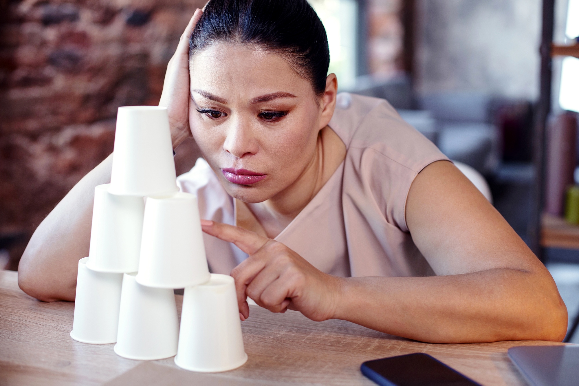 A woman rests her head on one hand while intently stacking paper cups into a pyramid on a desk. She appears focused, with a laptop and phone nearby. The setting is a mix of casual and work environment.