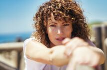 A woman with curly hair gazes into the camera with a serene expression. She is outdoors on a sunny day, resting her arms on a railing, with a blurry background of blue sky and water.