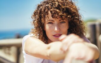 A woman with curly hair gazes into the camera with a serene expression. She is outdoors on a sunny day, resting her arms on a railing, with a blurry background of blue sky and water.