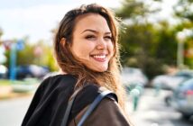 A woman with long hair smiles warmly while looking back at the camera. She is outdoors on a sunny day, with a blurred background of trees and parked cars.