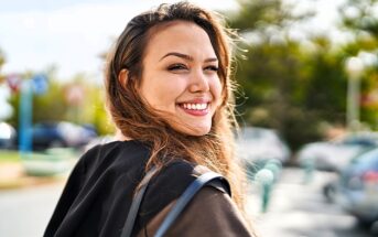 A woman with long hair smiles warmly while looking back at the camera. She is outdoors on a sunny day, with a blurred background of trees and parked cars.