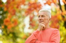 An older man with gray hair stands outdoors, wearing a coral sweater. He is looking thoughtfully into the distance, with one hand resting on his chin. The background features vibrant orange and green foliage, indicating a fall setting.