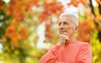 An older man with gray hair stands outdoors, wearing a coral sweater. He is looking thoughtfully into the distance, with one hand resting on his chin. The background features vibrant orange and green foliage, indicating a fall setting.