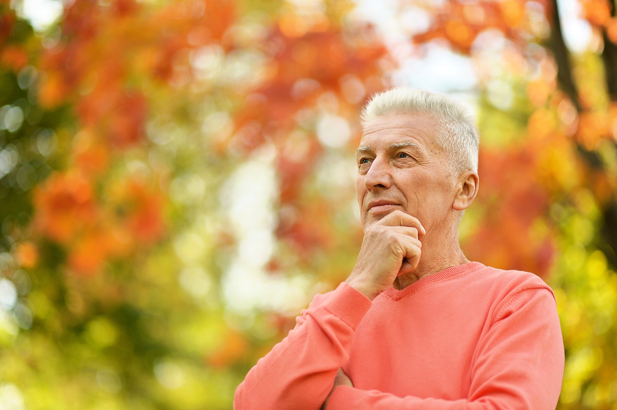 An older man with gray hair stands outdoors, wearing a coral sweater. He is looking thoughtfully into the distance, with one hand resting on his chin. The background features vibrant orange and green foliage, indicating a fall setting.