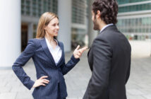 A woman and a man in business suits are standing outdoors and engaging in a conversation. The woman gestures with her hand while smiling. They appear to be in a modern urban setting with glass buildings in the background.