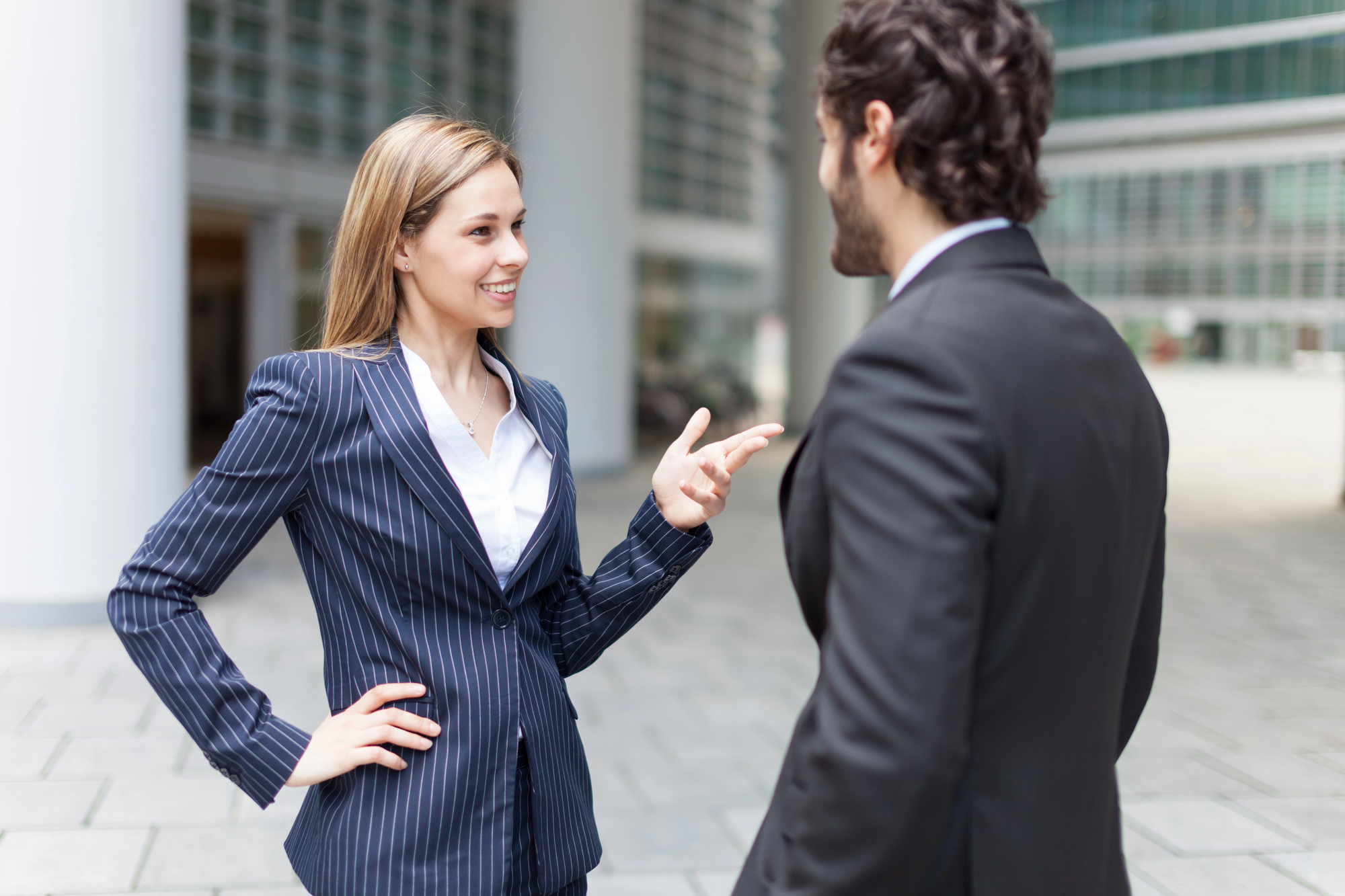 A woman and a man in business suits are standing outdoors and engaging in a conversation. The woman gestures with her hand while smiling. They appear to be in a modern urban setting with glass buildings in the background.