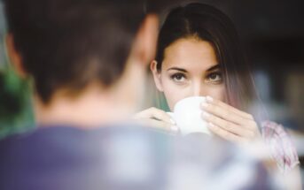 A woman with long brown hair looks over the rim of a white cup she is holding, appearing thoughtful. The blurred silhouette of another person is in the foreground, suggesting a conversation or shared moment.